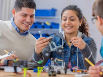 Cheerful female high school student enjoys building a robot in technology class. Classmates and a teacher are helping her. 